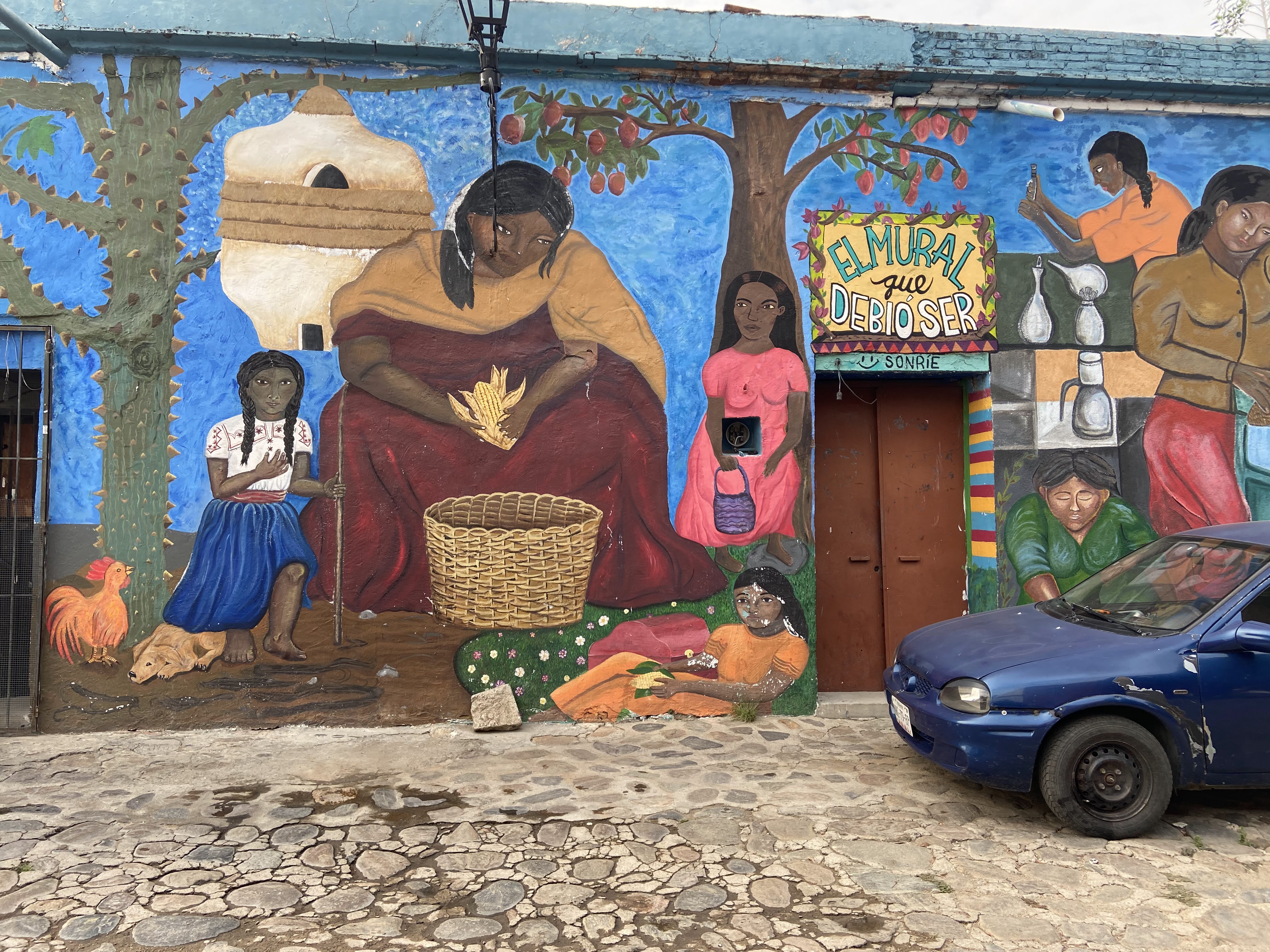 Photo of Mural in Oaxaca of people working in the community.  Blue car in front. 
